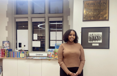 Samia Hersi standing in front of a bookshelf, surrounded by various books of different sizes and colors.